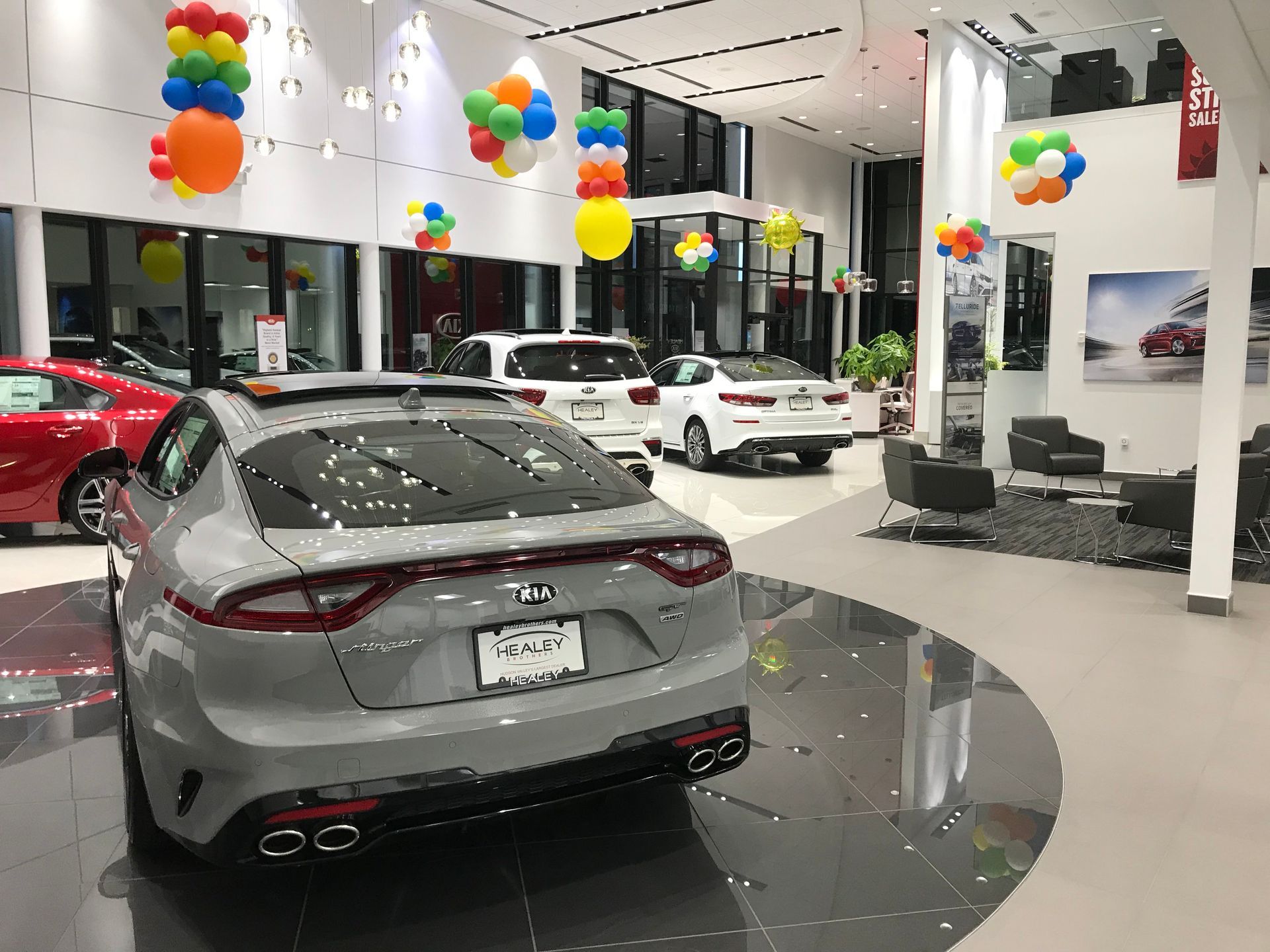 A grey Kia Stinger displayed in a brightly lit car dealership showroom decorated with colorful balloon clusters.