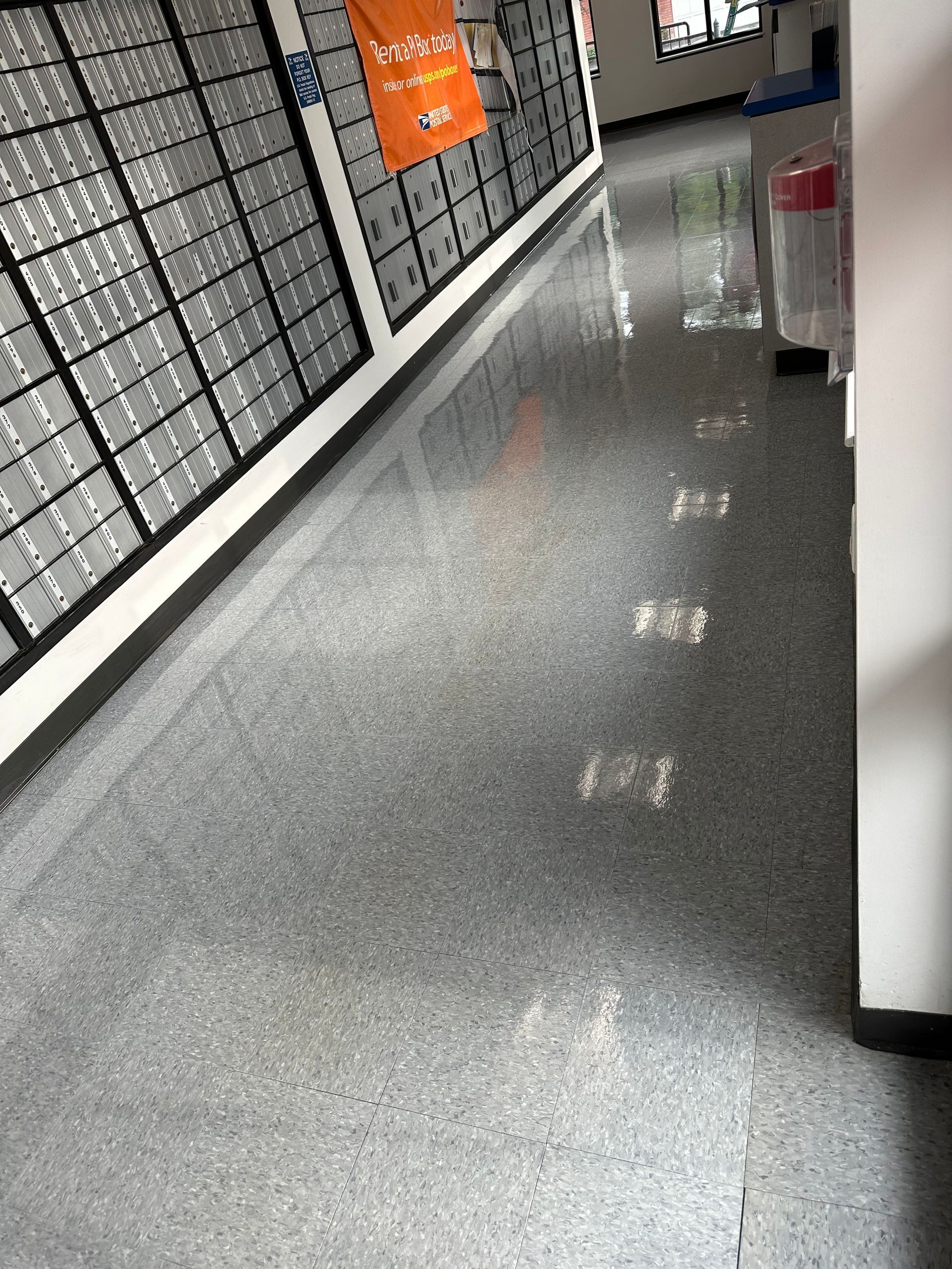 An indoor hallway with a long wall of mailboxes on the left, leading to a windowed doorway.