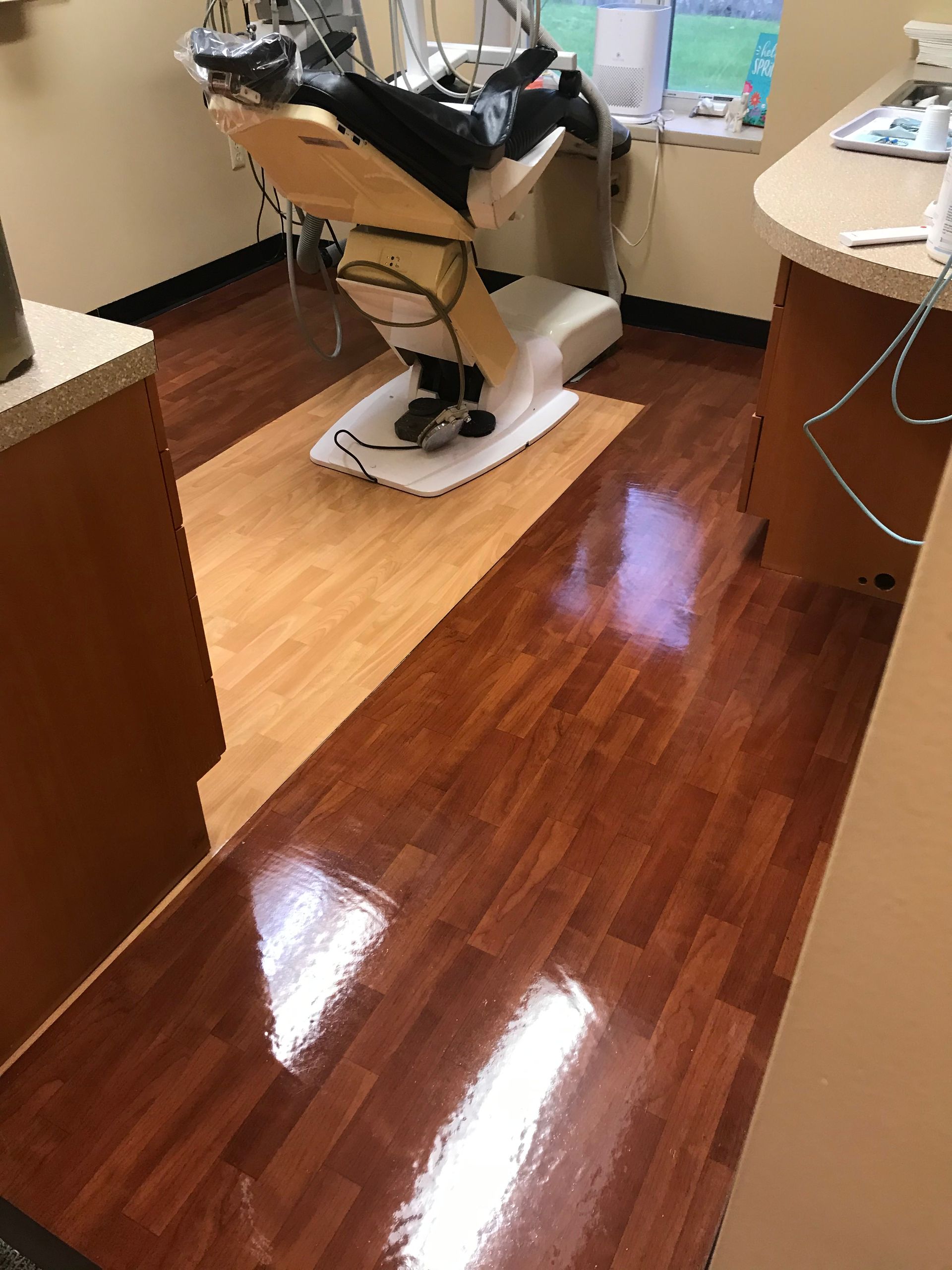 An empty dental exam room featuring a dental chair, light-colored wood flooring under the chair, and polished floor tiles.