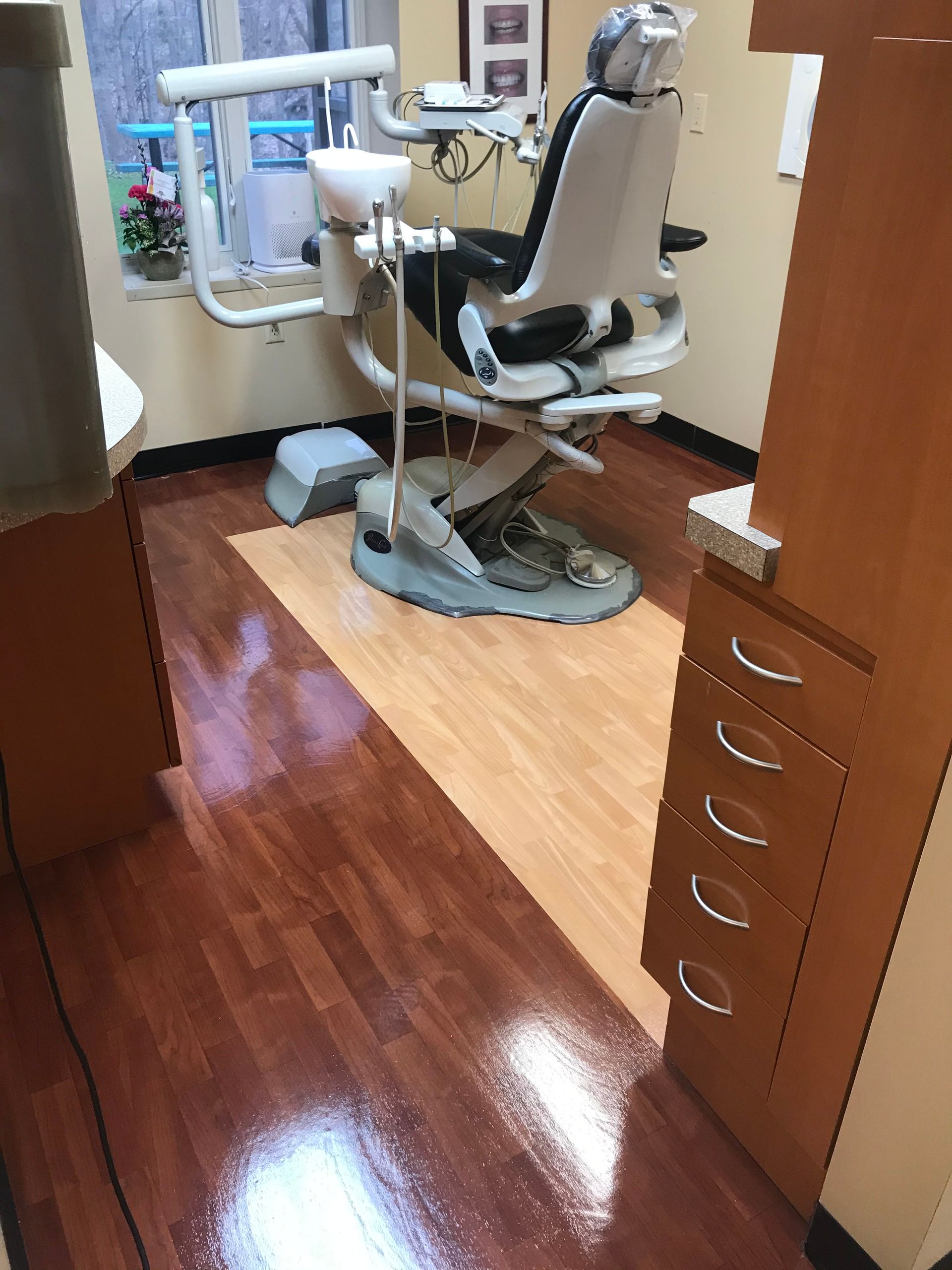 A dental exam room with a reclining chair, equipment stand, and light fixture, featuring light and dark wood flooring.