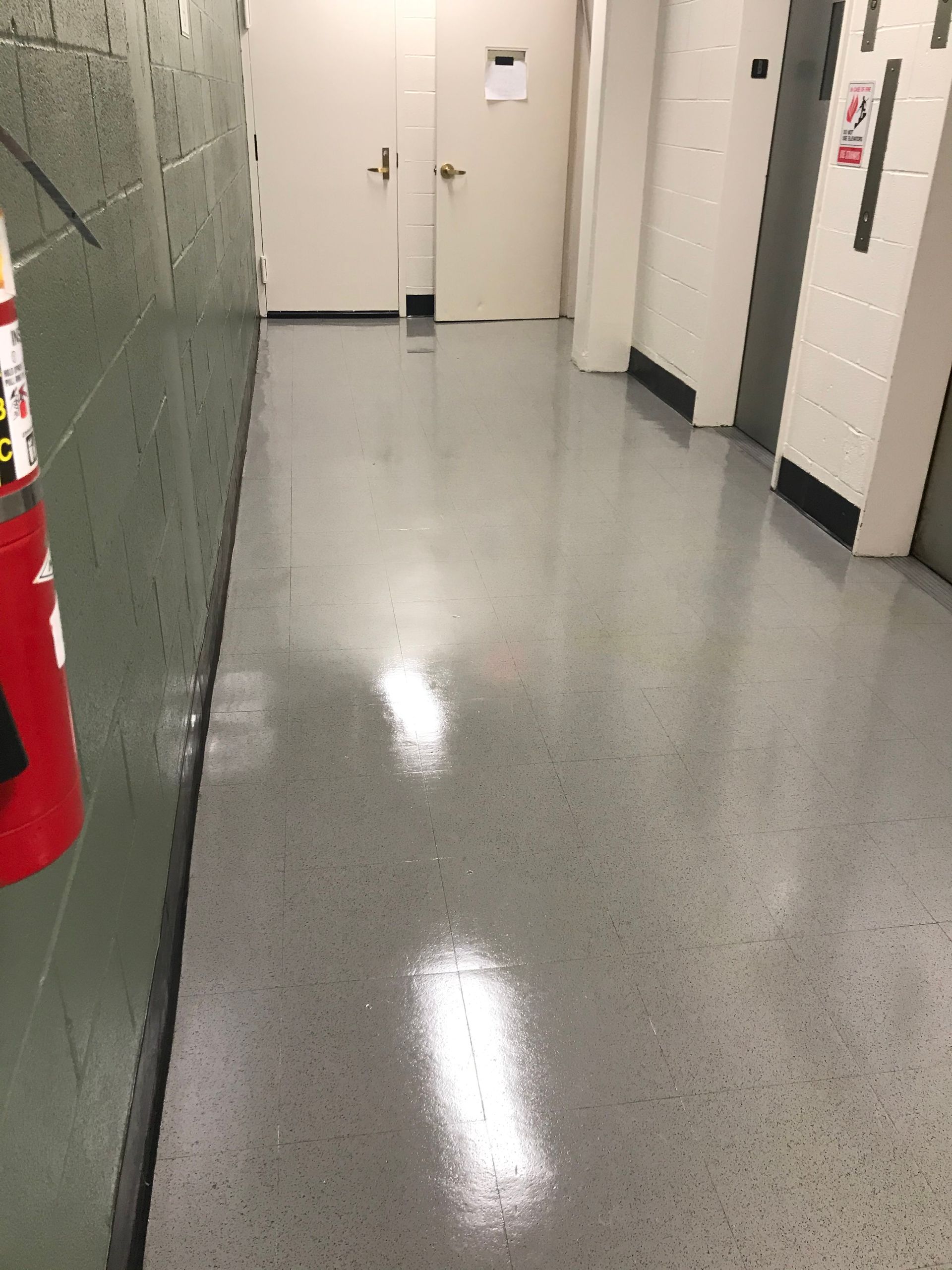 A view down a corridor with shiny gray epoxy flooring, white doors, and a red fire extinguisher on a green wall.