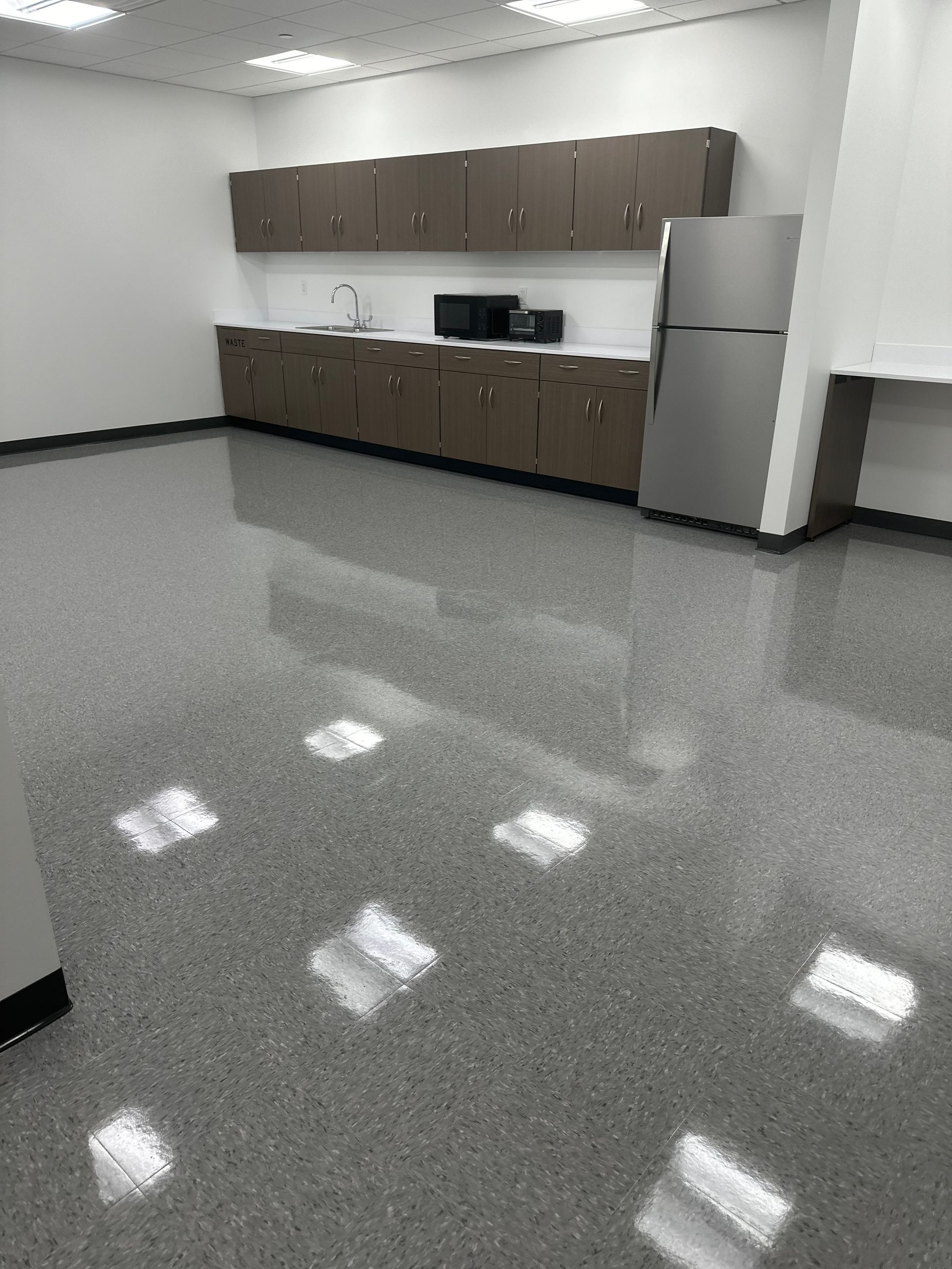A brightly lit, modern office kitchenette with brown cabinets, a stainless steel refrigerator, and a glossy gray floor.