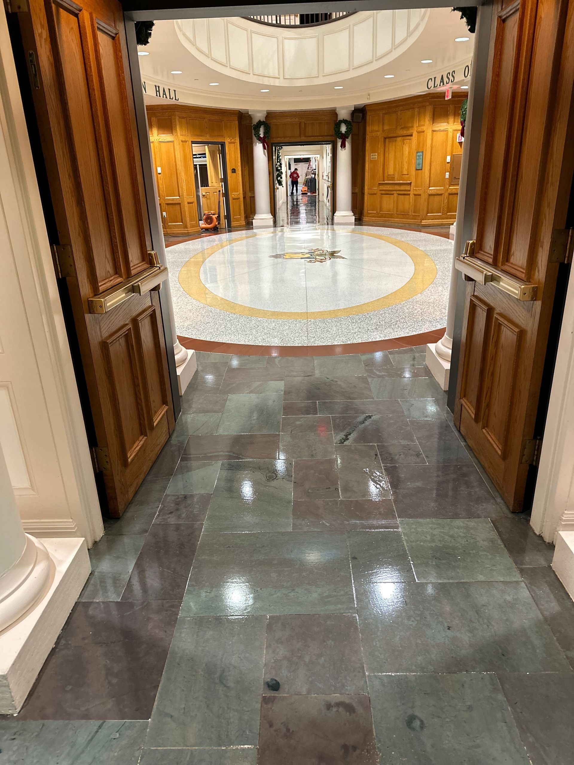 Double wooden doors open into a bright, circular lobby with wood-paneled walls and a patterned tile floor.