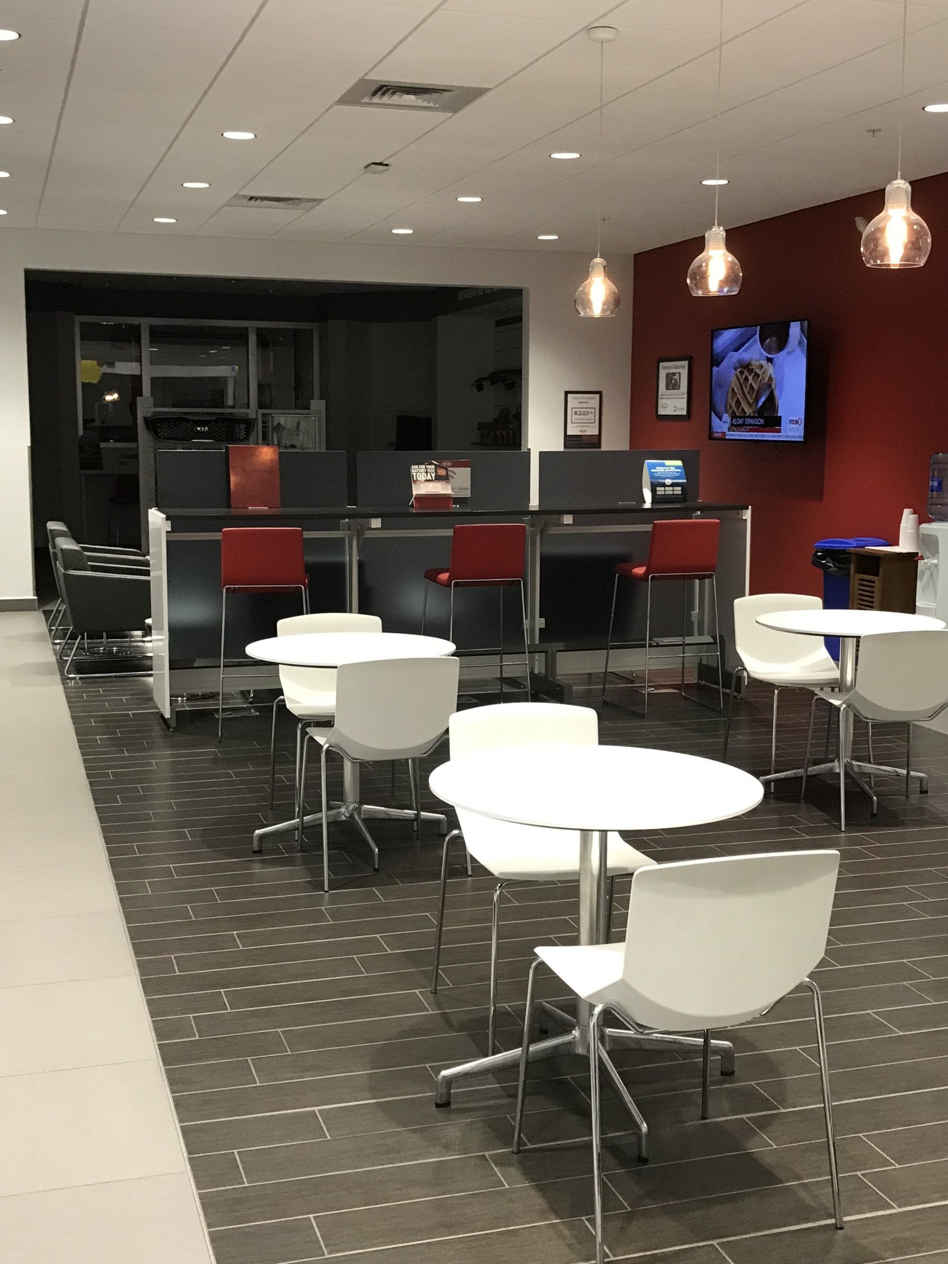 Modern office break room with white tables and chairs, bar seating at a counter, and a TV mounted on a red wall.