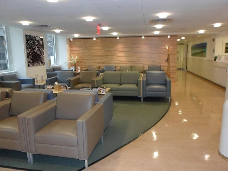 Modern waiting room with beige leather chairs, green sofas, a wood-paneled wall, and a curved green rug on tiled flooring.