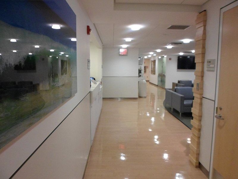 A bright, modern hospital hallway with polished floors, a painting on the left wall, and waiting area chairs on the right.
