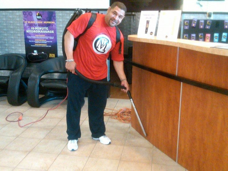 A person in a red t-shirt uses a backpack vacuum to clean the base of a wooden counter in a brightly lit office.