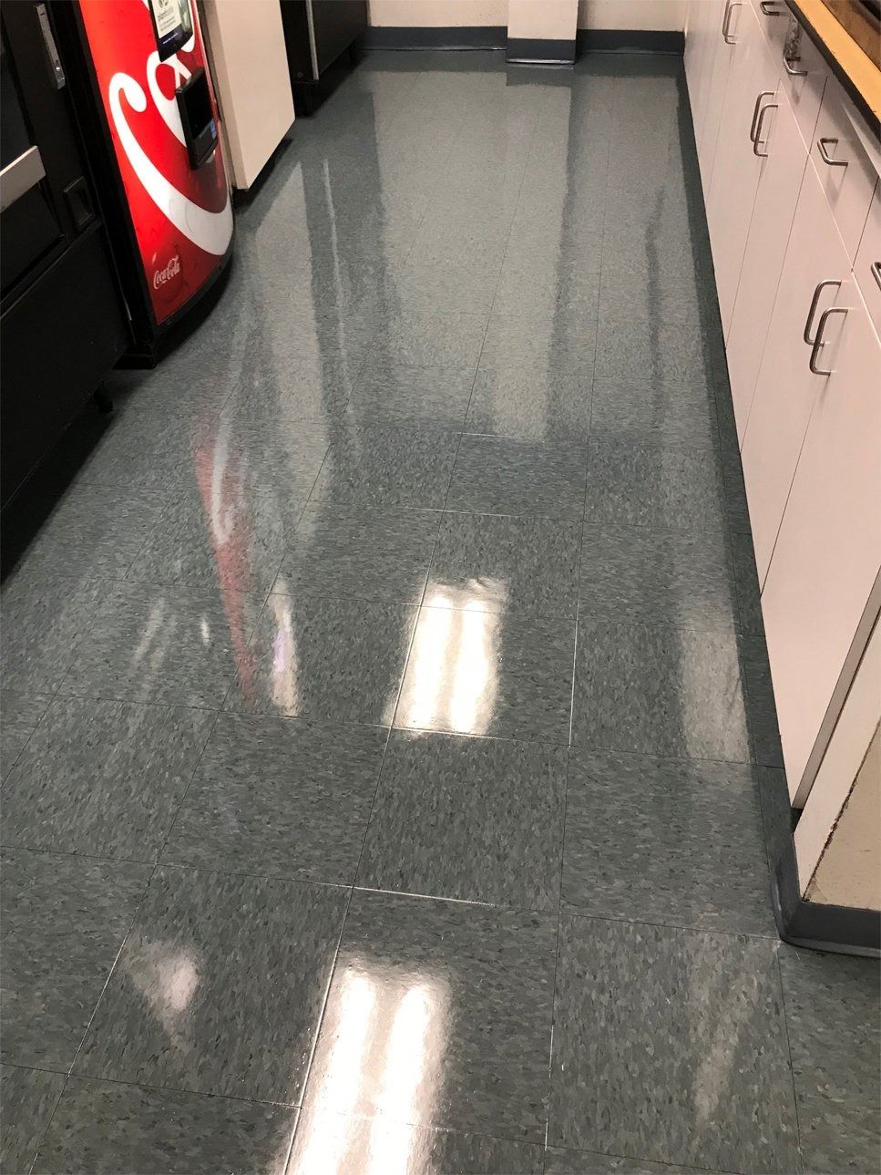 A brightly lit, clean, and highly polished dark gray speckled floor in a hallway with white cabinets and a vending machine.