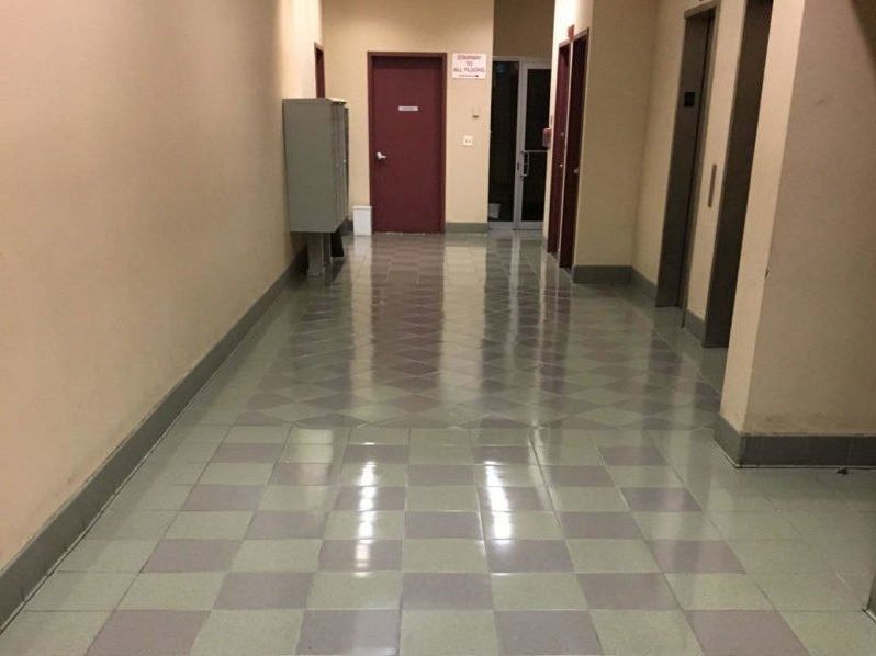 A brightly lit, empty hallway with light and dark square floor tiles, beige walls, and several dark-colored doors.