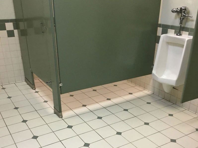 A urinal mounted on a tiled wall next to a public restroom stall with a olive green privacy partition.