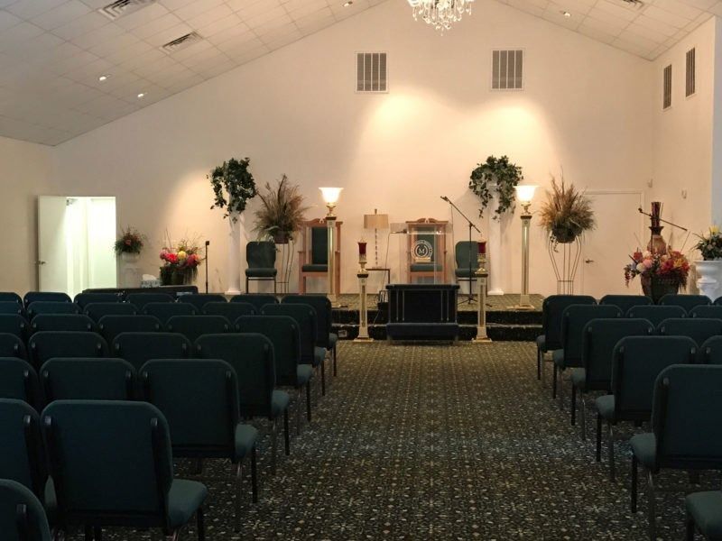 An empty church interior with rows of dark blue chairs facing a carpeted stage featuring floral arrangements and a podium.