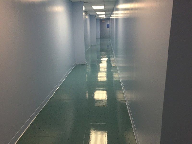 A long, empty indoor hallway with light-colored walls and a glossy, reflective dark green floor illuminated by overhead lights.