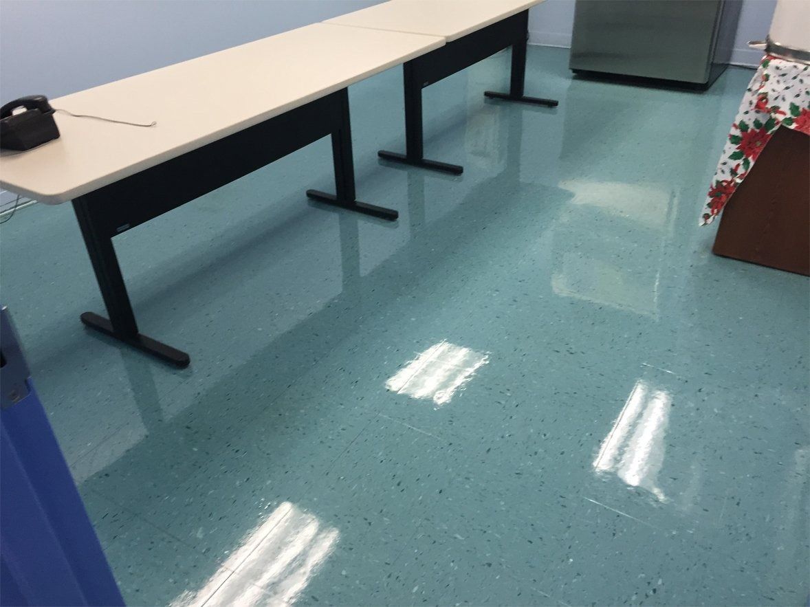 A pale blue office room featuring two long, light-colored tables on T-shaped black legs atop a glossy, speckled floor.