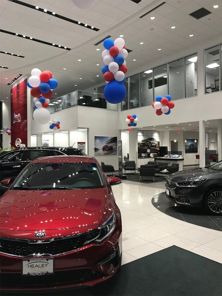 A shiny red sedan displayed in a bright, modern car dealership decorated with clusters of red, white, and blue balloons.