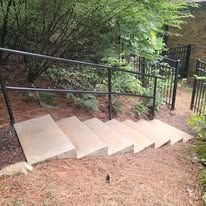 Concrete steps with black handrails leading up a hill, surrounded by foliage.