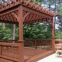 Wooden pergola with deck, surrounded by railing, set in a lush outdoor area.