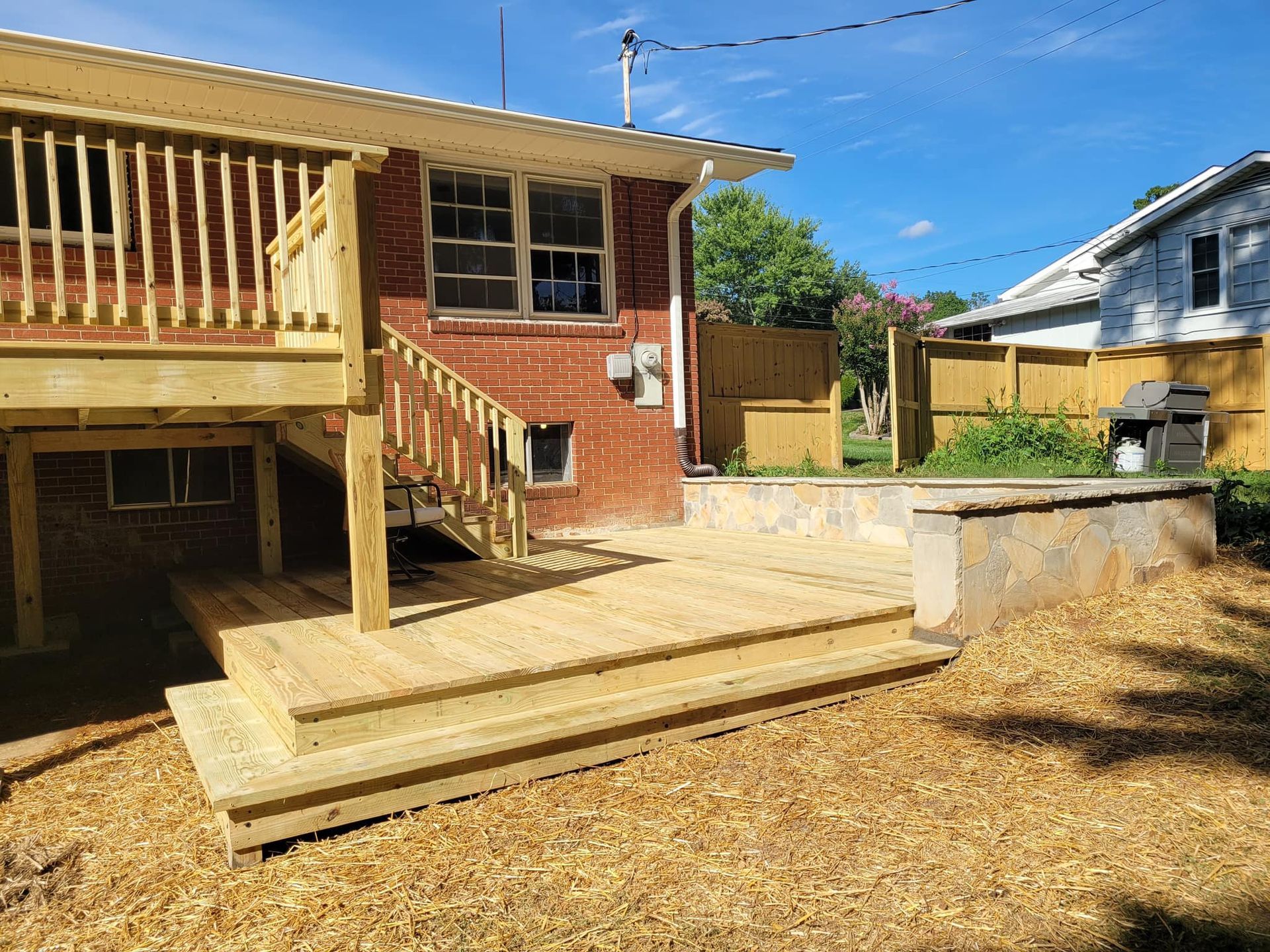 Wooden deck with steps in a backyard next to a brick house and a retaining wall, sunny day.