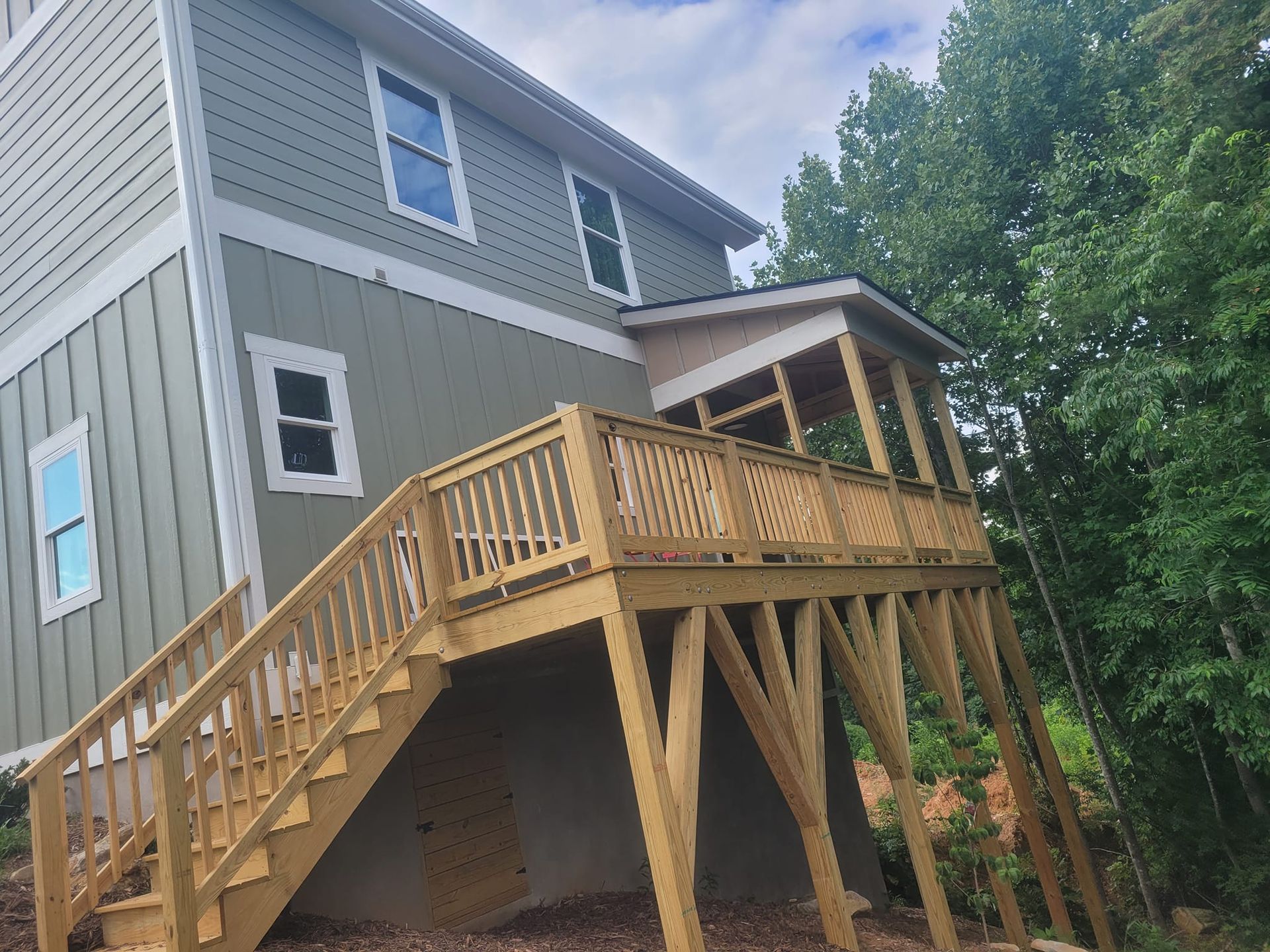 Two-story house with a wooden deck, stairs, and a screened porch, next to green trees.