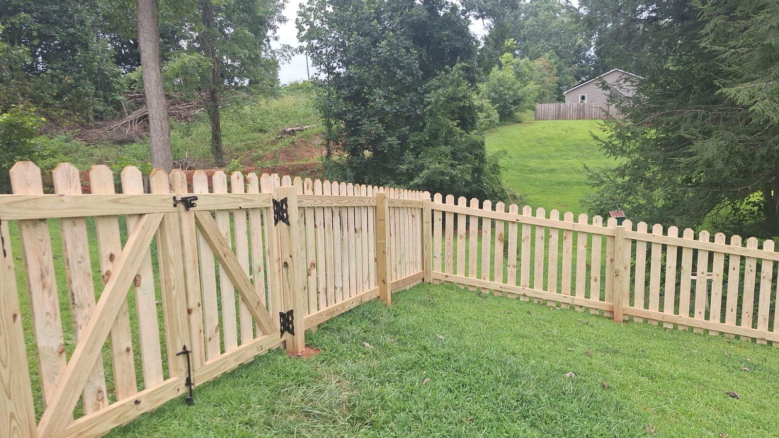 A wooden picket fence with a gate in a grassy backyard, surrounded by trees.