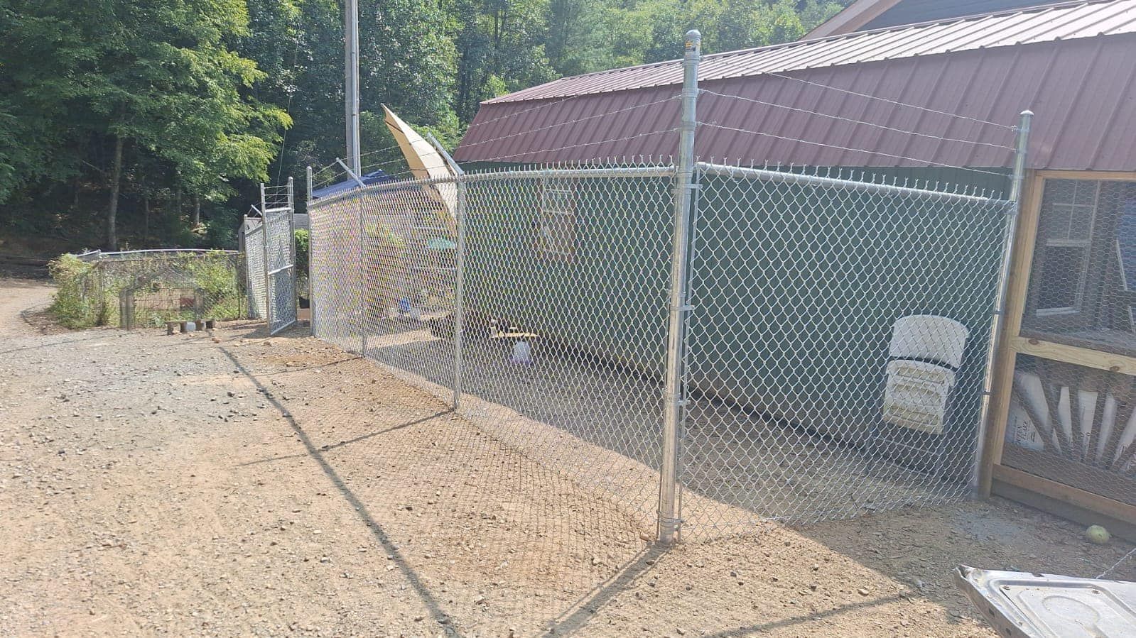 Chain-link fence around a gravel area next to a building with a red roof and trees in the background.