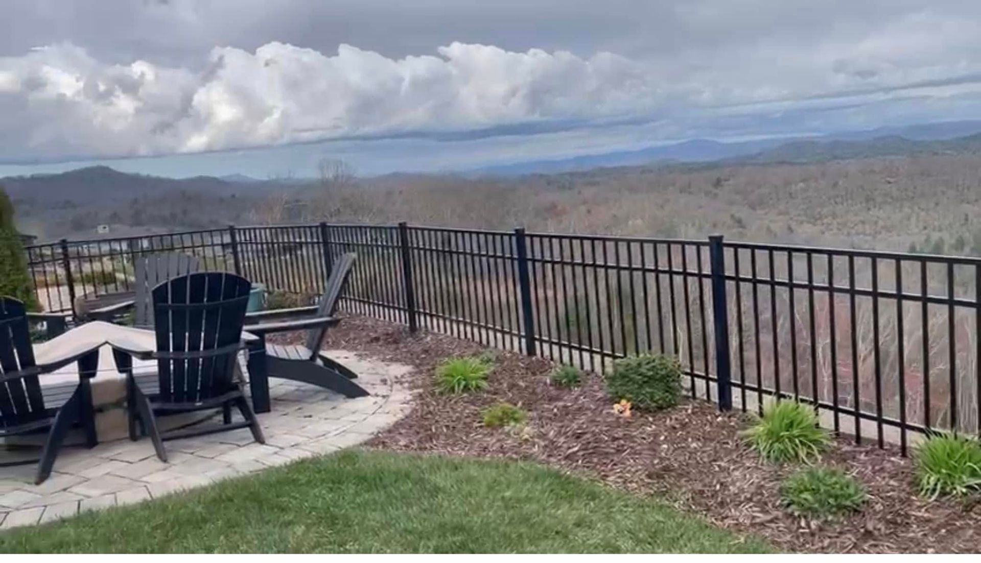 Black fence surrounds a patio with Adirondack chairs overlooking a mountain view under a cloudy sky.