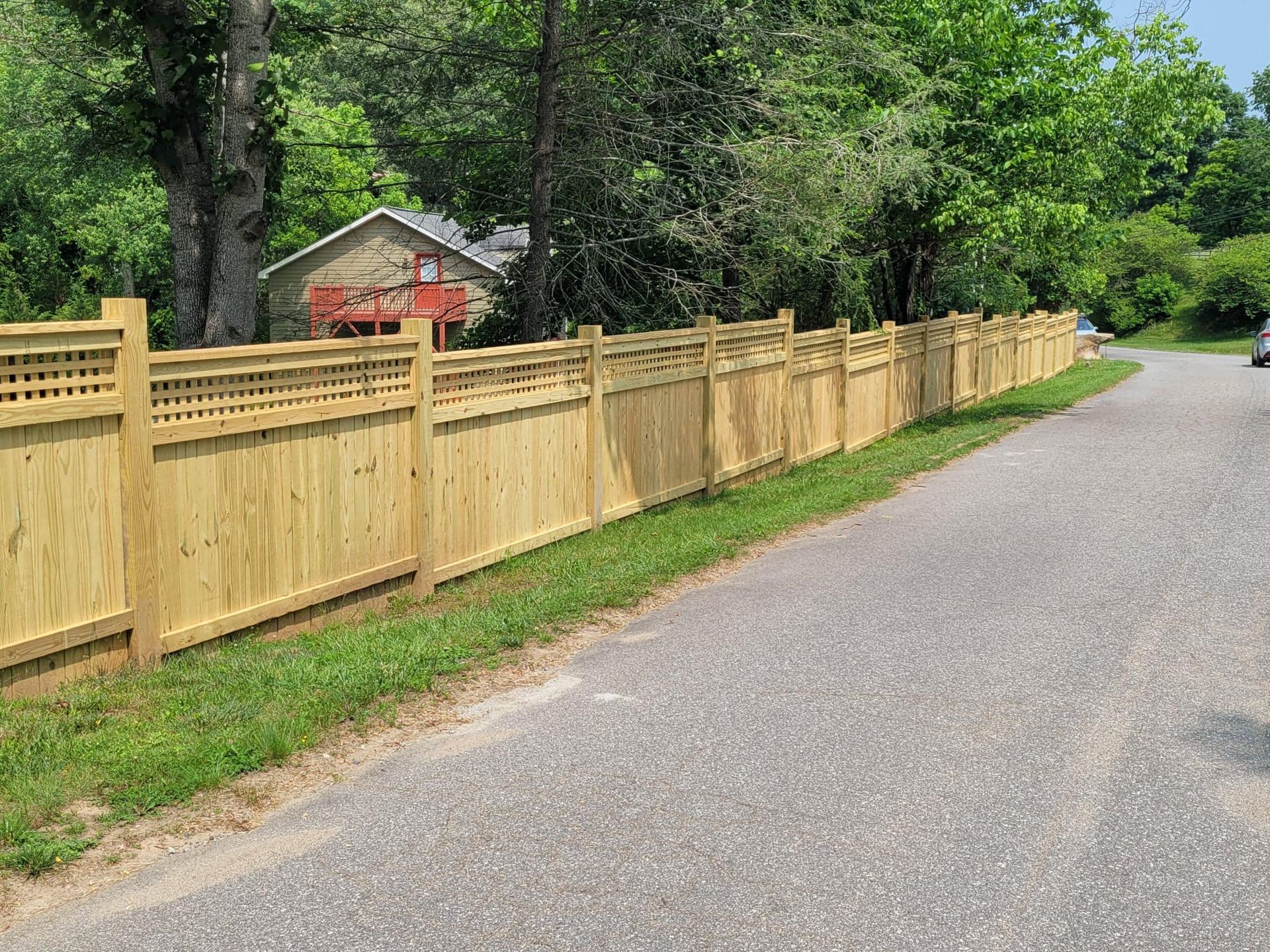 Wooden fence along a paved road, with a house and trees in the background.