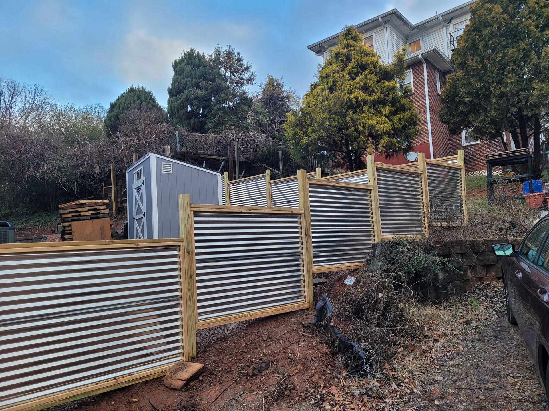 Corrugated metal and wood fence bordering a garden area, with a shed and trees in the background.
