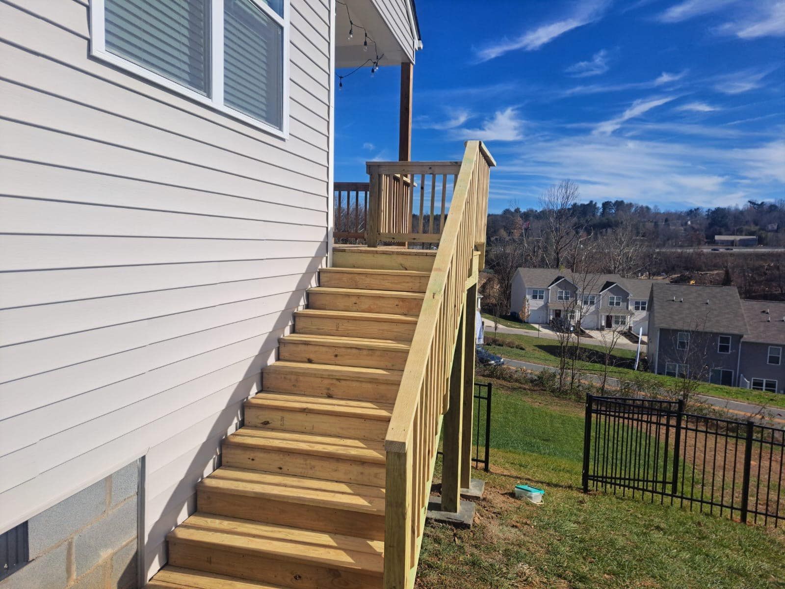 Wooden outdoor staircase leading to a deck with a house on the left and a green yard with houses in the distance.