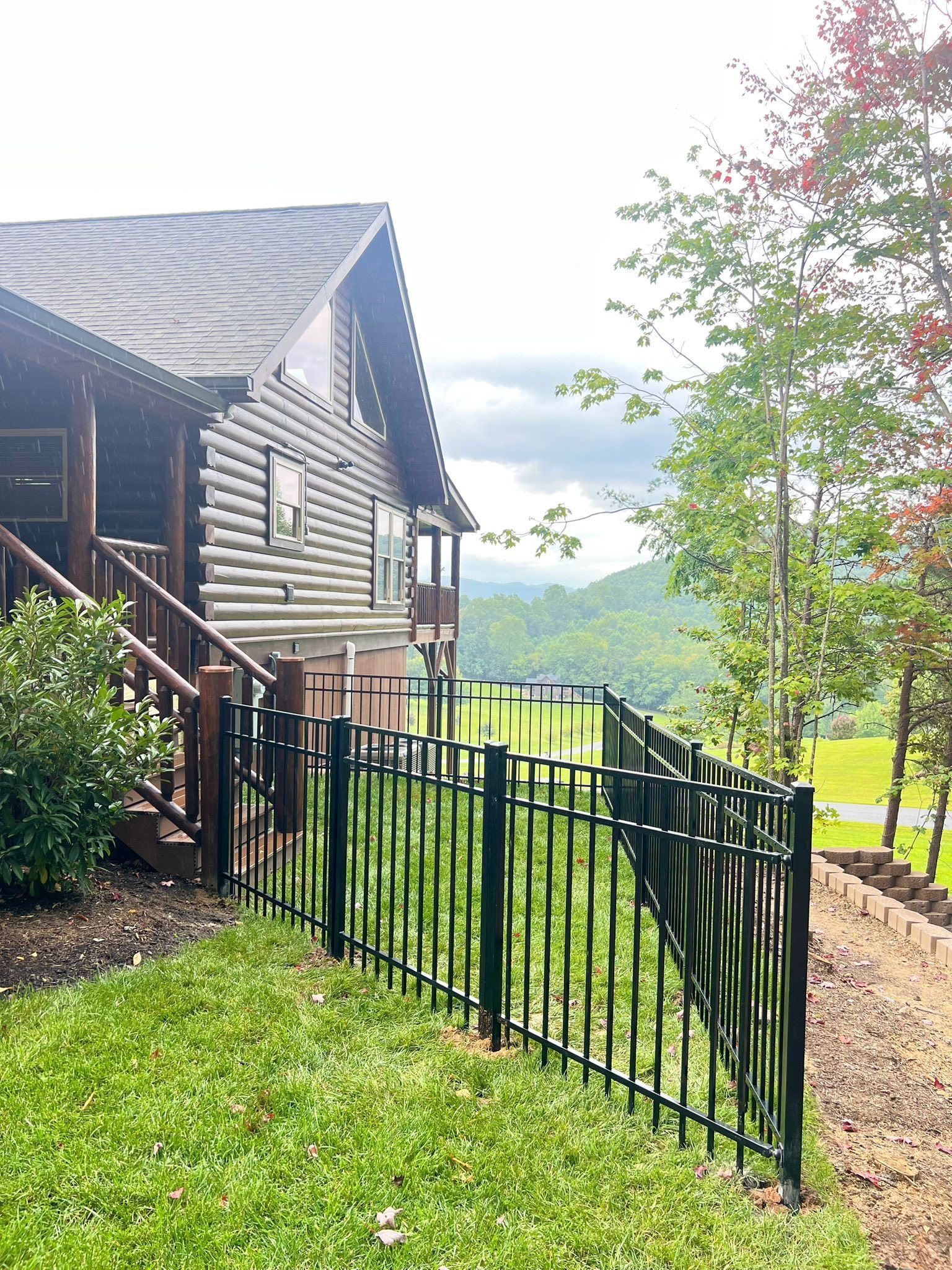 Wooden cabin with black fence overlooking a valley. Green grass and trees in the background.