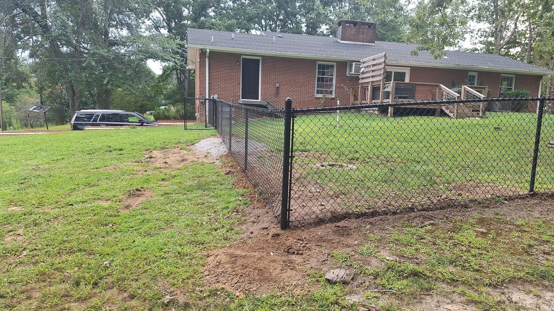Black chain-link fence around a grassy yard in front of a brick house. A car is in the background.