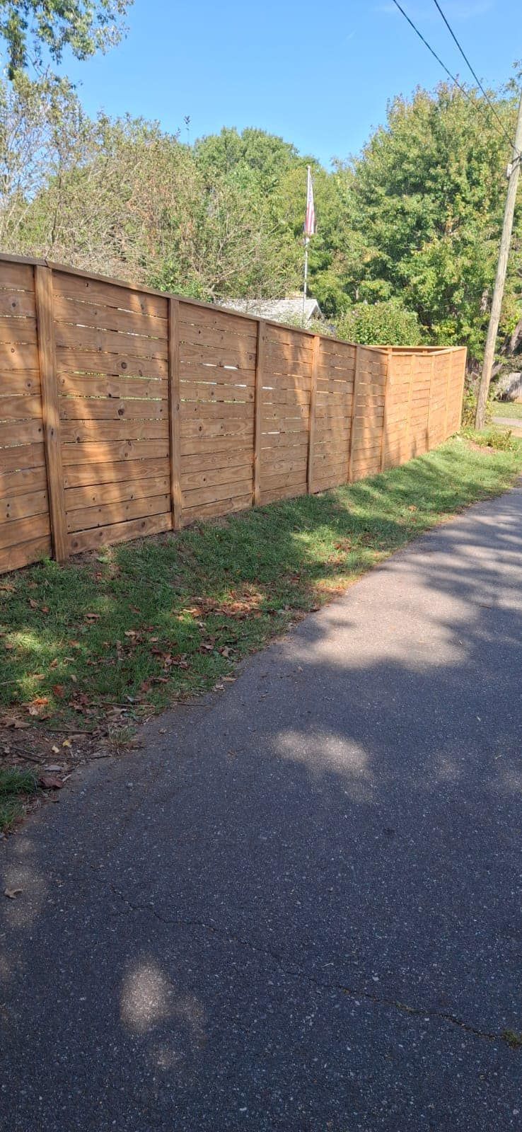 Brown brick wall alongside a gray asphalt road, green grass, and trees on a sunny day.