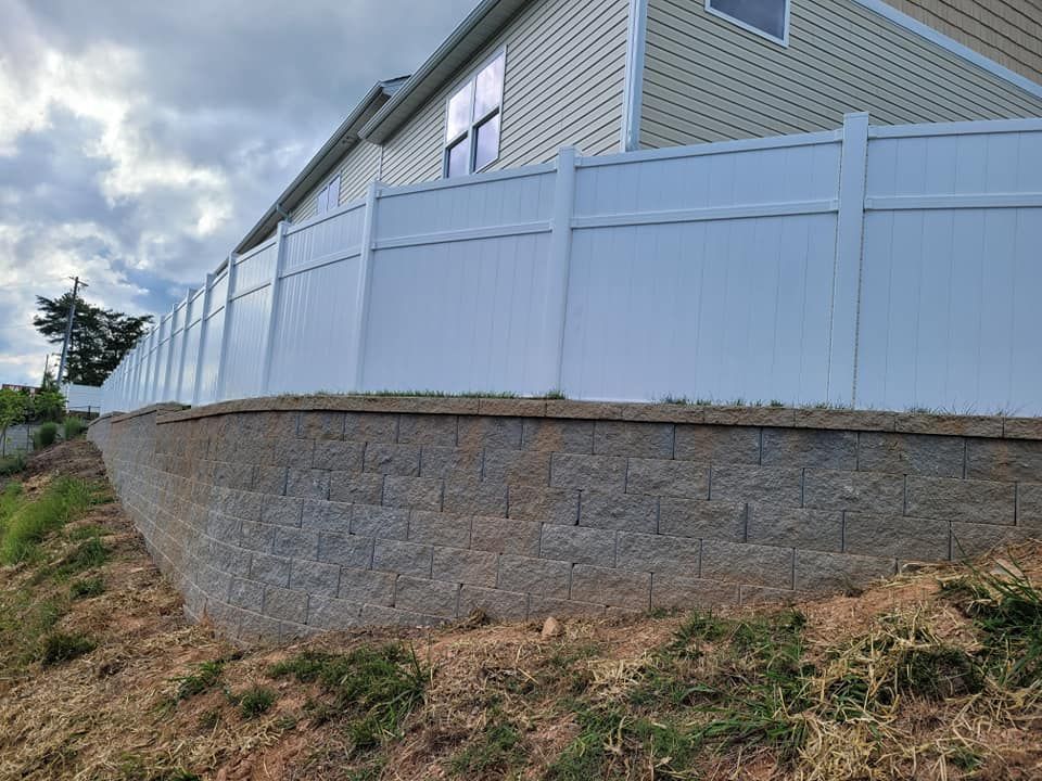 White vinyl fence atop a retaining wall made of gray blocks, on a grassy hillside.