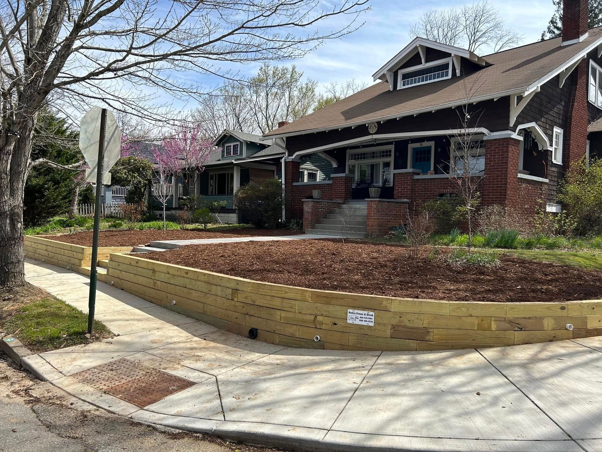 Wooden retaining wall in front of a house with wood chips and a sidewalk on a sunny day.