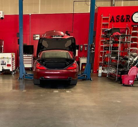 Red SUV in a mechanic’s garage with its hood open and trunk raised, flanked by blue lifts and shelves of parts