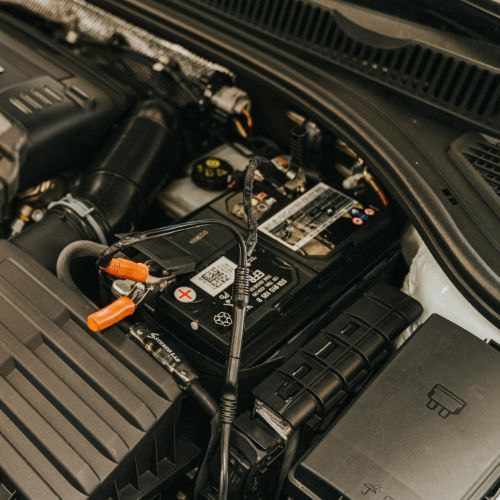 Car battery in an engine bay with jumper cables attached and orange clamps.