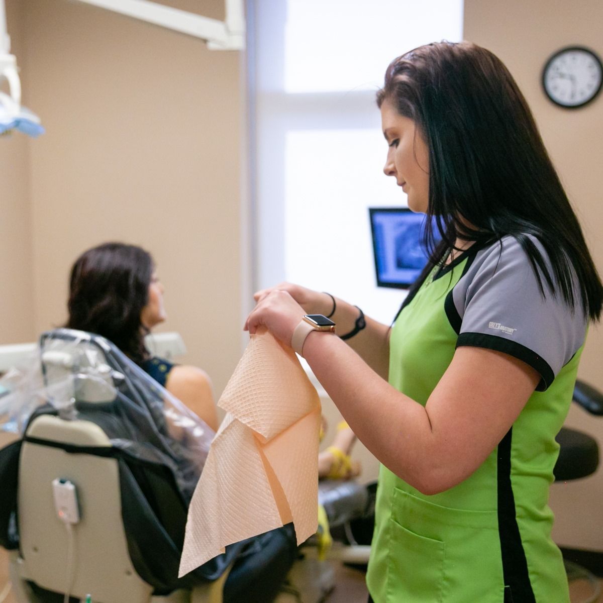 Dental assistant preparing a patient for treatment, holding paper bib. Patient sits in dental chair.