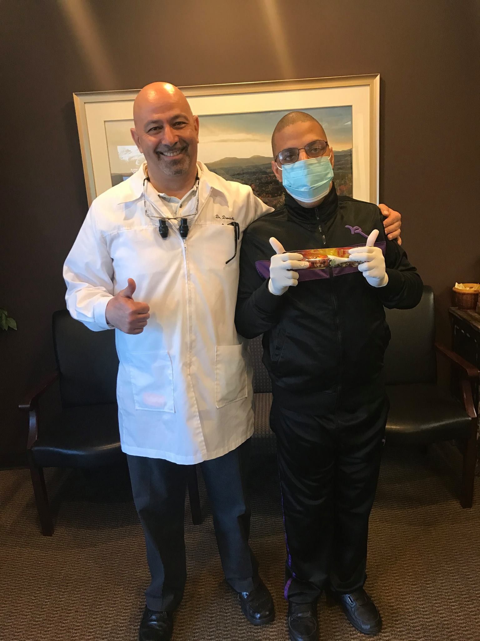 Two men smiling, one in a lab coat, other in track suit wearing mask and gloves, holding dental work.