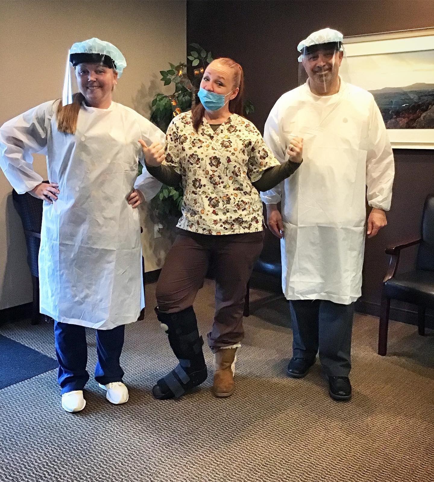 Three people pose in a room. Two wear surgical gowns and caps. The center person wears scrubs, a mask, and boots.