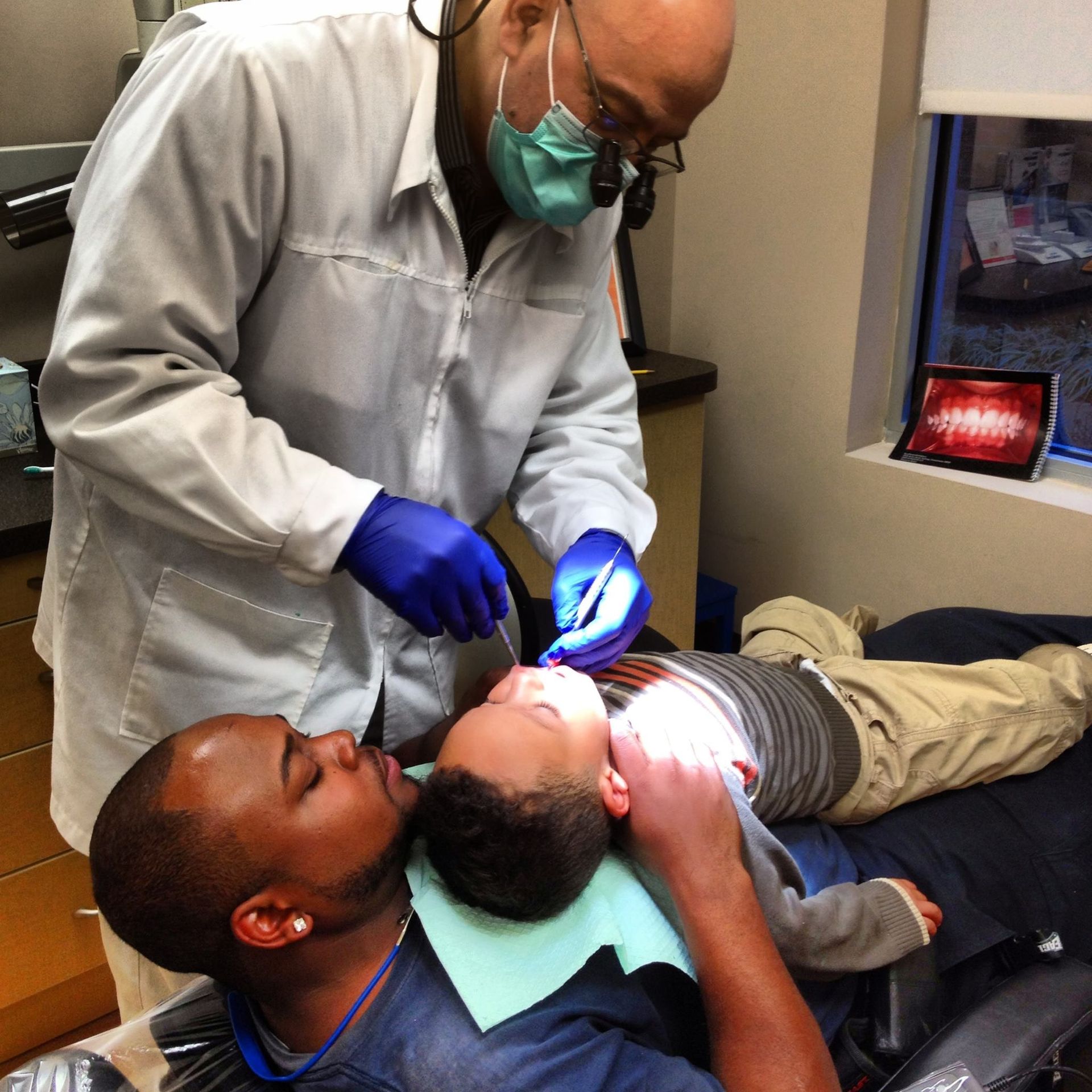 Dentist examining a child's teeth while the child's father holds the child. Dentist is in a white lab coat and gloves.