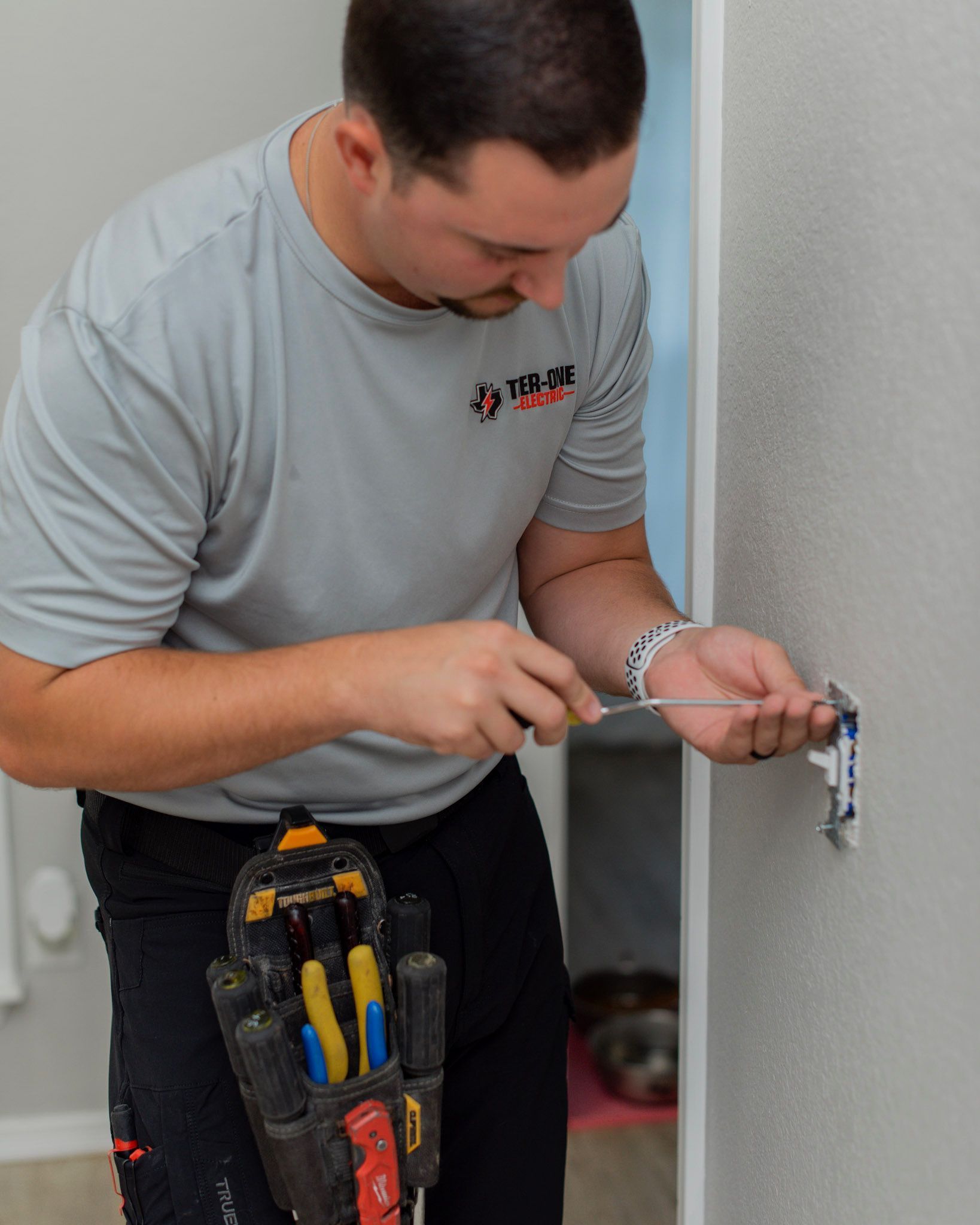 Person in a gray shirt using tools to work on a wall outlet in a room