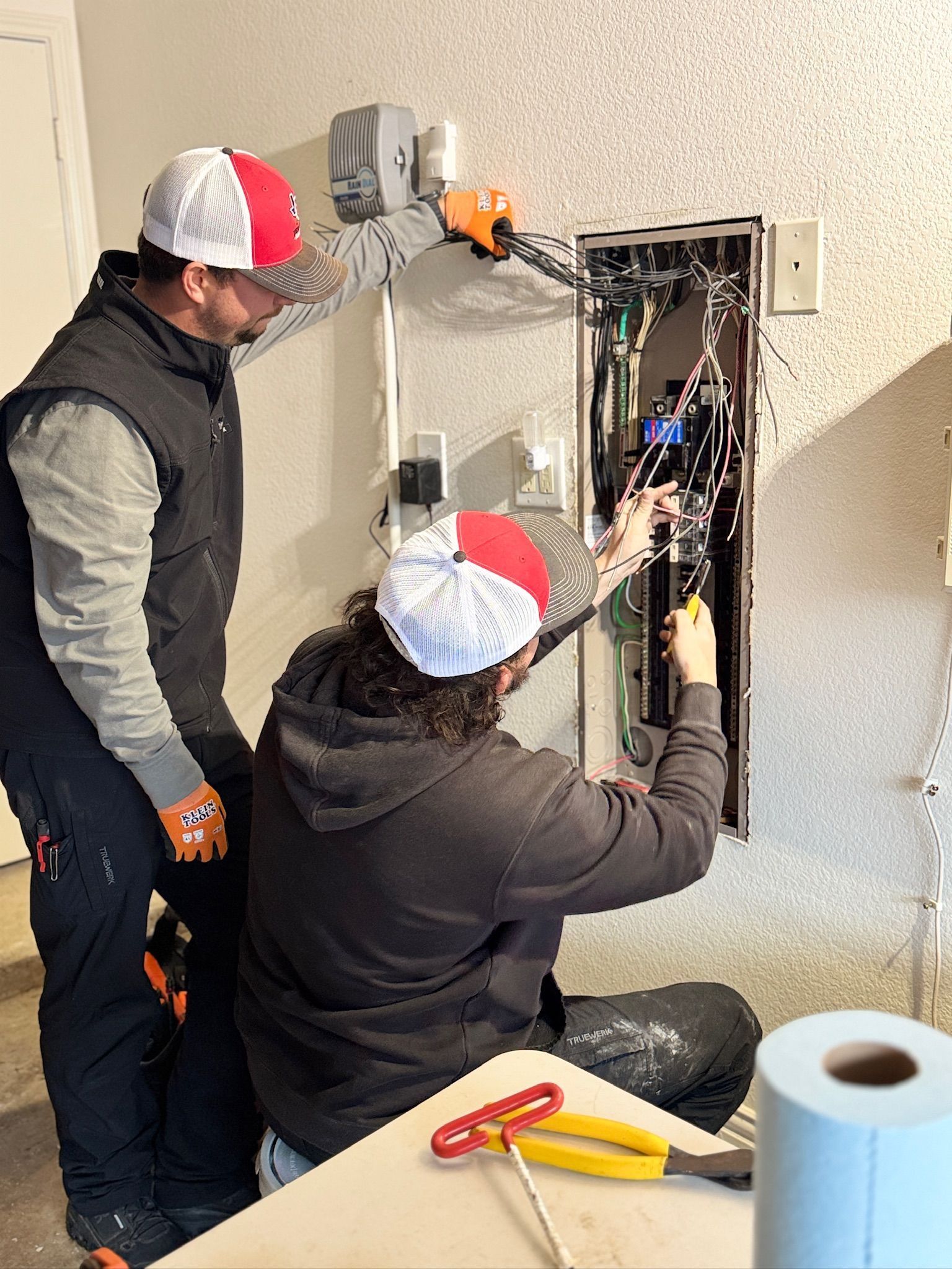 Two technicians wiring an open electrical panel in a workshop, one standing and one kneeling with tools.