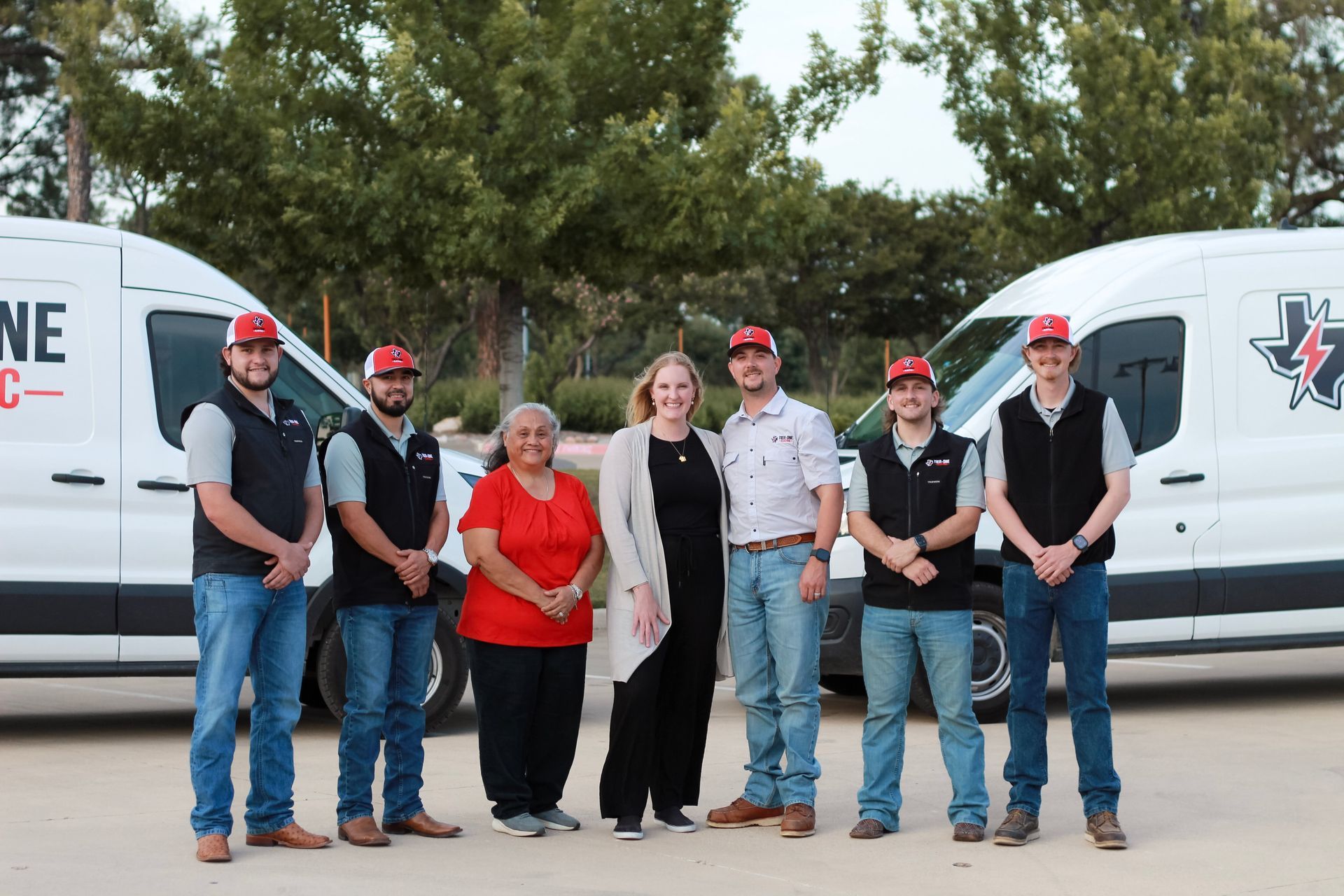 Seven people posing between two white service vans outdoors, all wearing work shirts and caps.