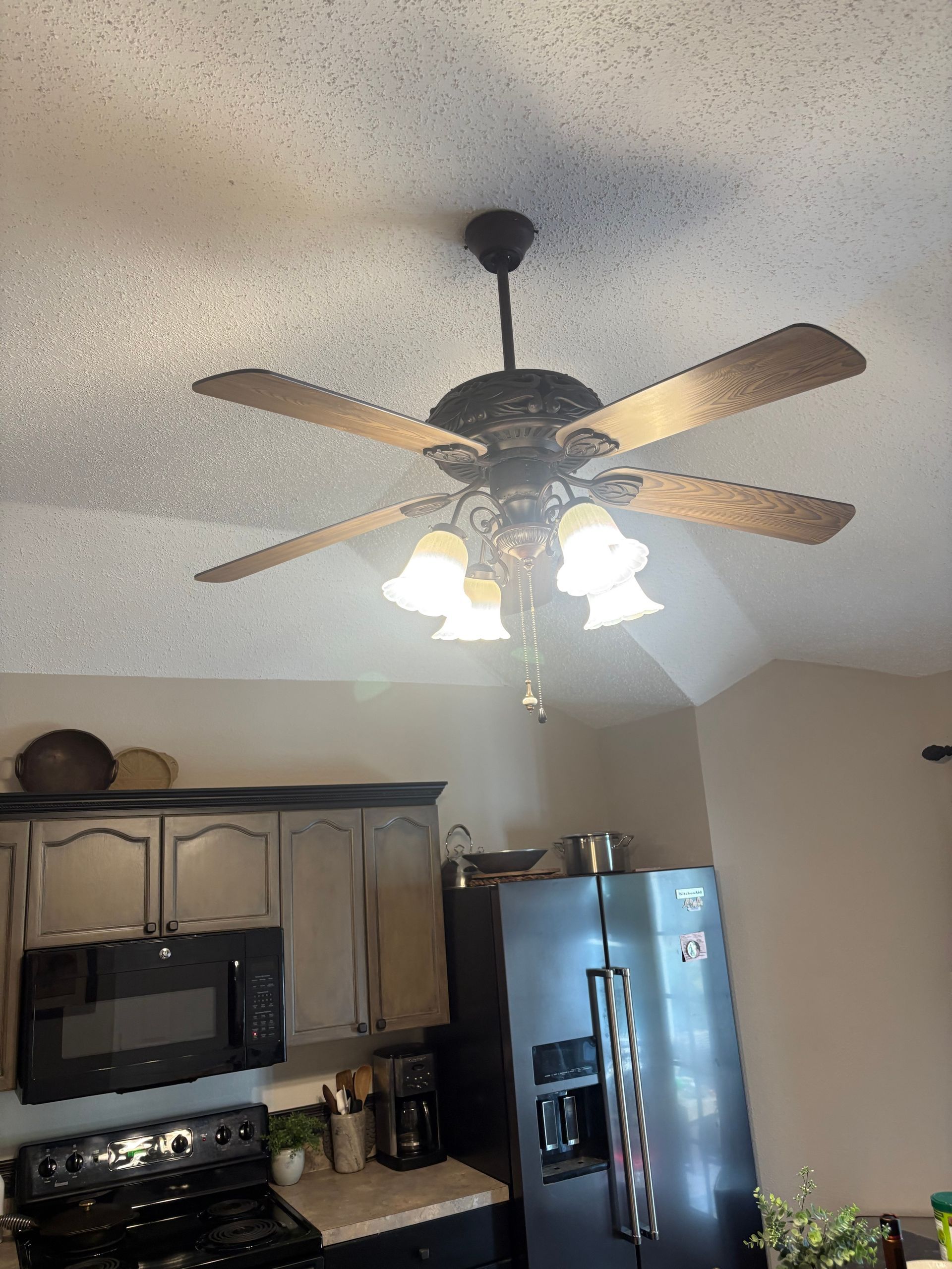Kitchen with dark cabinets, stainless fridge, and a ceiling fan with lights on a speckled ceiling.