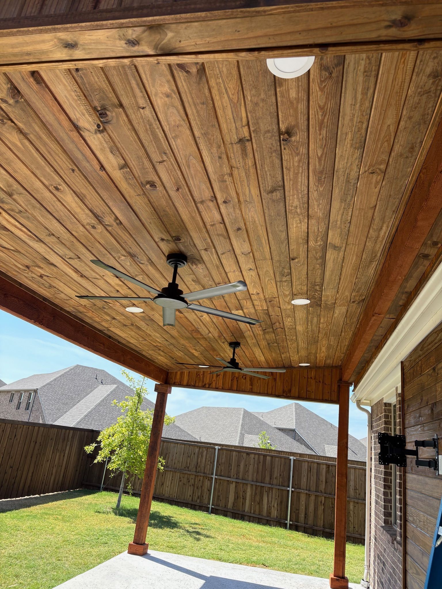 Covered backyard patio with wood ceiling, ceiling fan, brick wall, and fenced lawn behind.