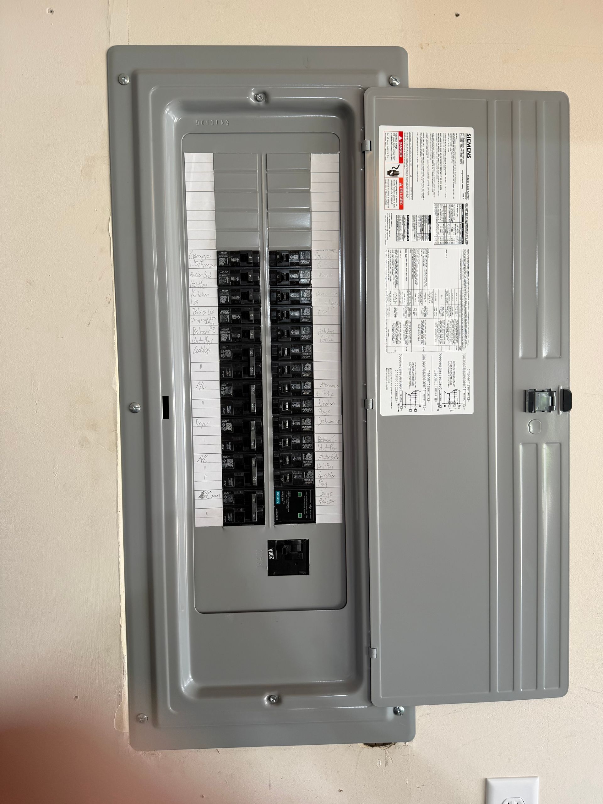 Open electrical breaker panel with gray cover and rows of black circuit breakers on a wall