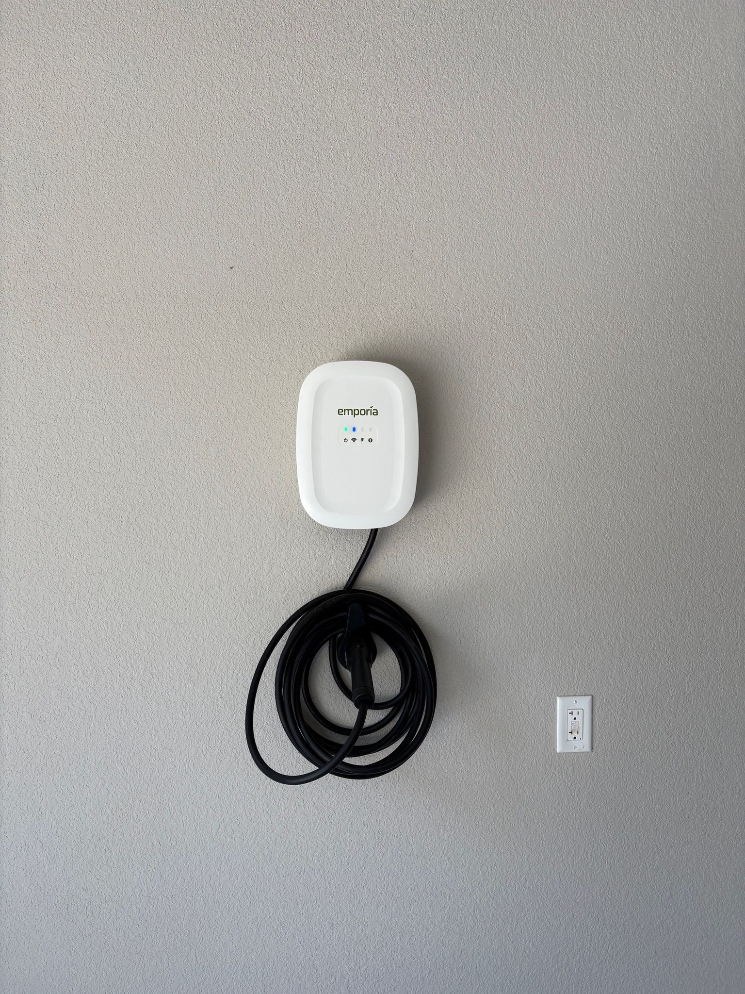 Wall-mounted white EV charger with coiled black cable on a gray wall.