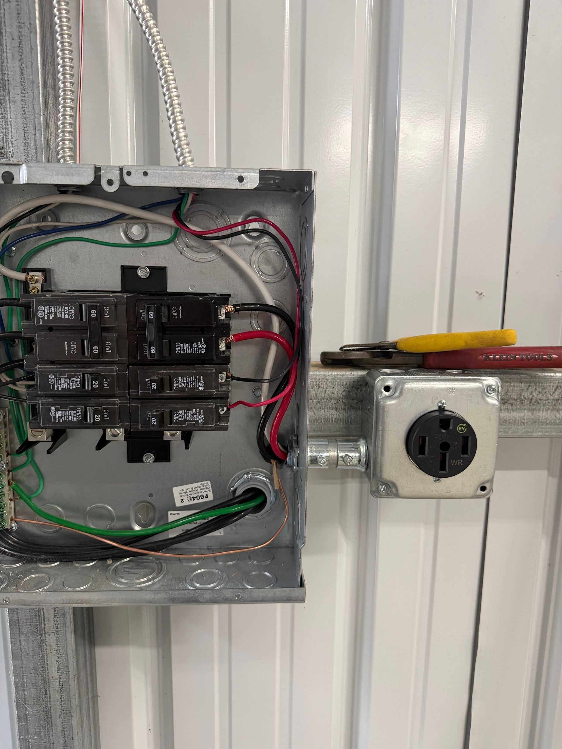 Open electrical panel with breakers and wiring beside a wall outlet box with a screwdriver on top