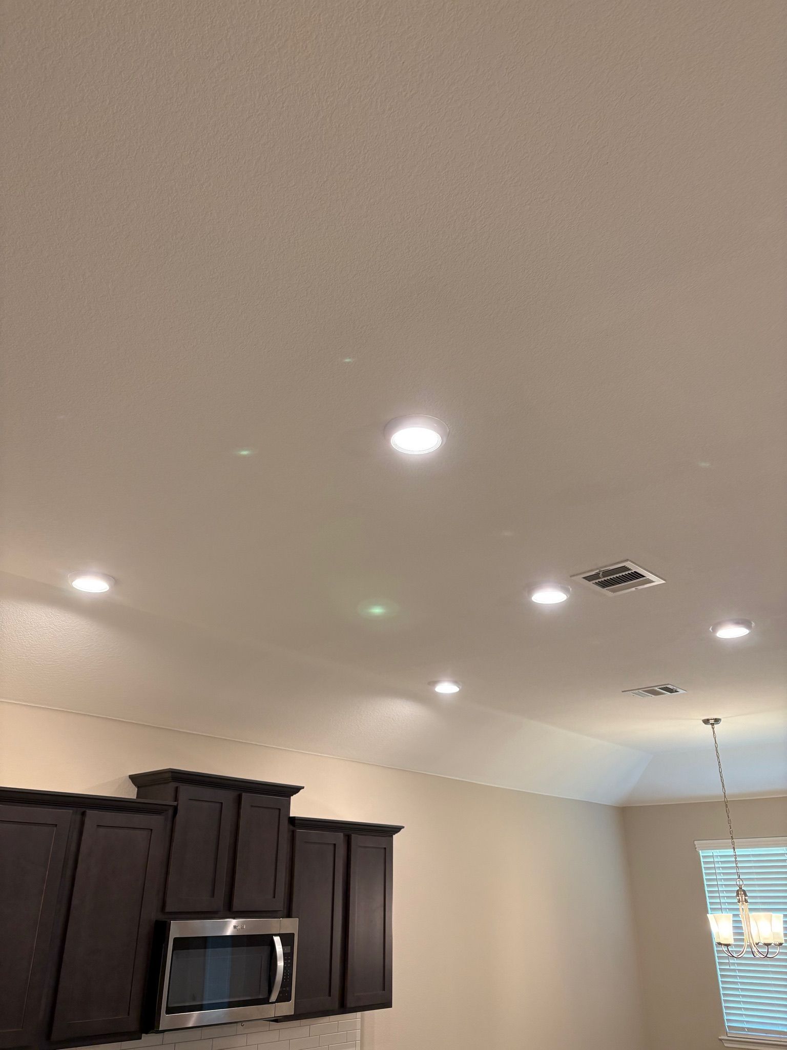 Kitchen ceiling with recessed lights above dark cabinets and a microwave, with a window in the background