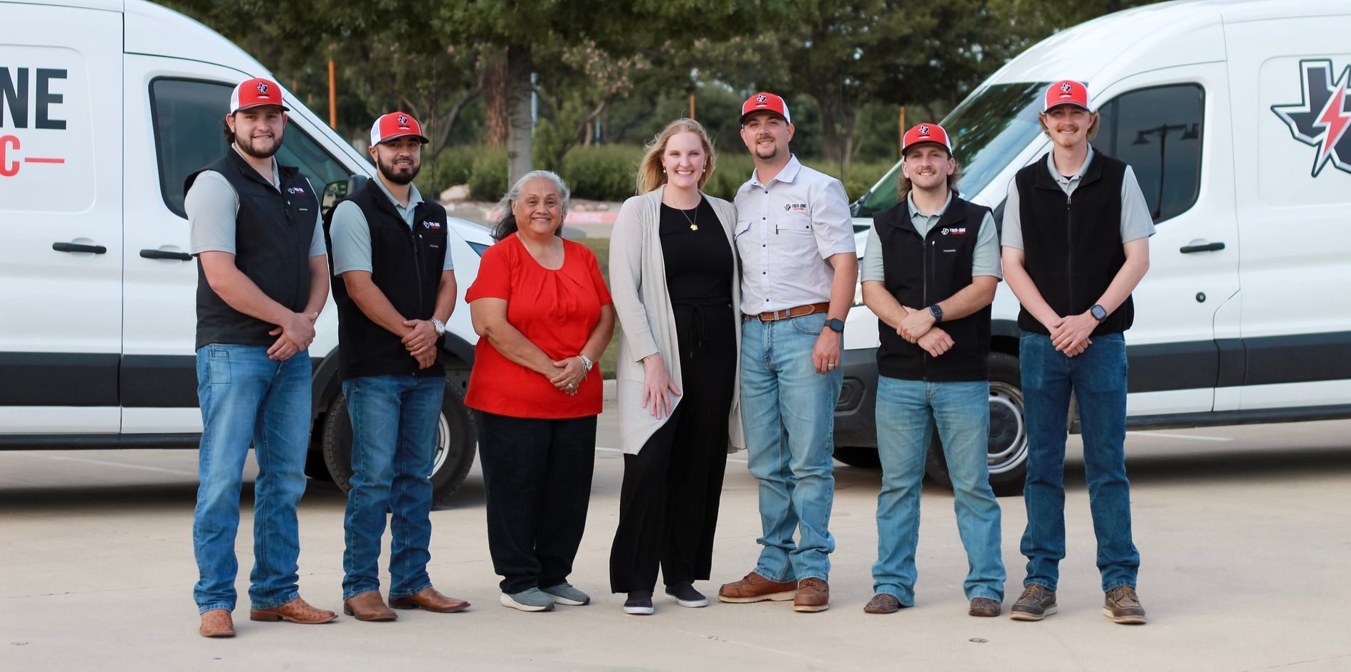 Six people in red caps and black shirts stand in front of white service vans outdoors.