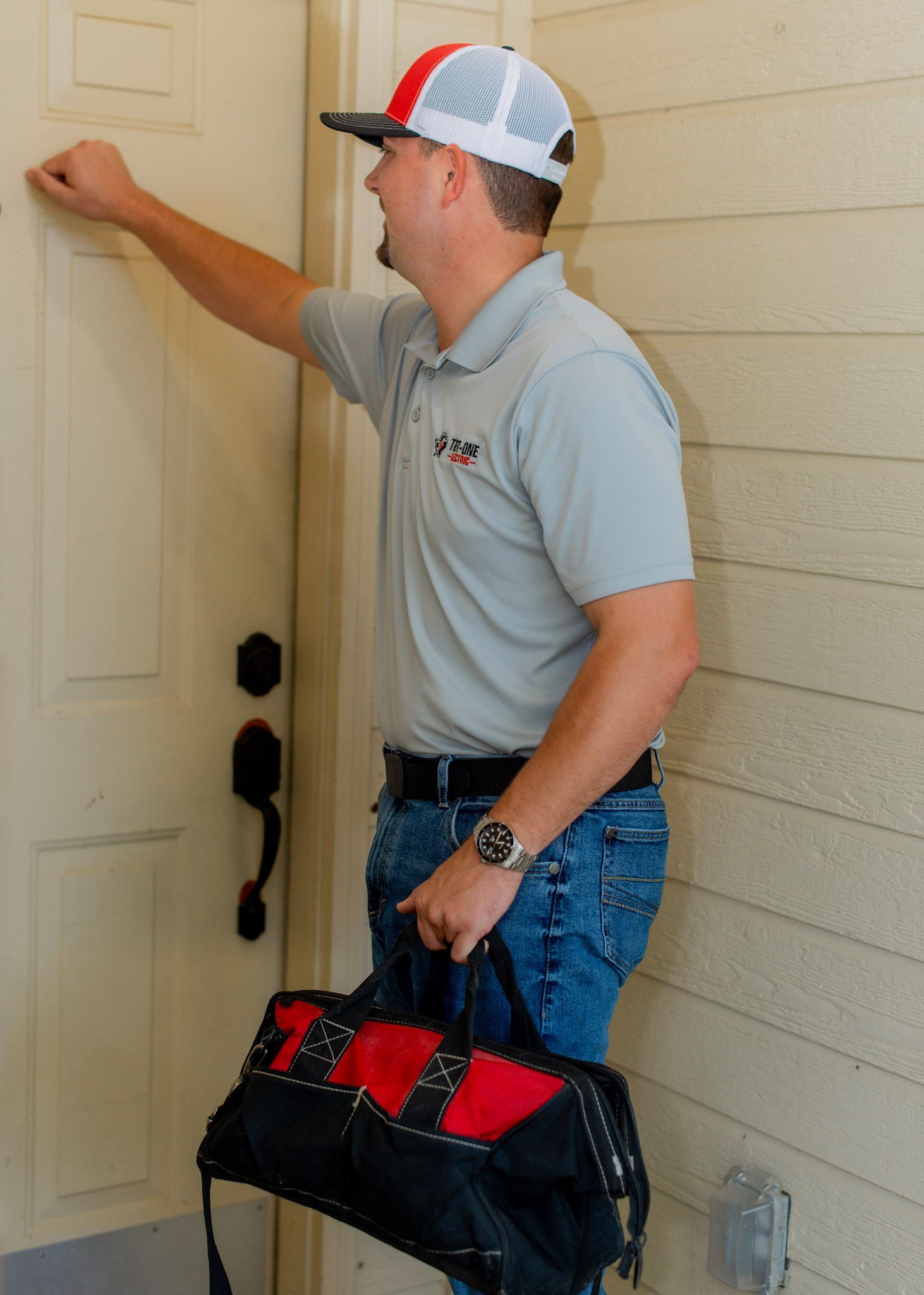 Man in work attire knocks on a door, holding a tool bag. He wears a hat and smiles, standing outside.
